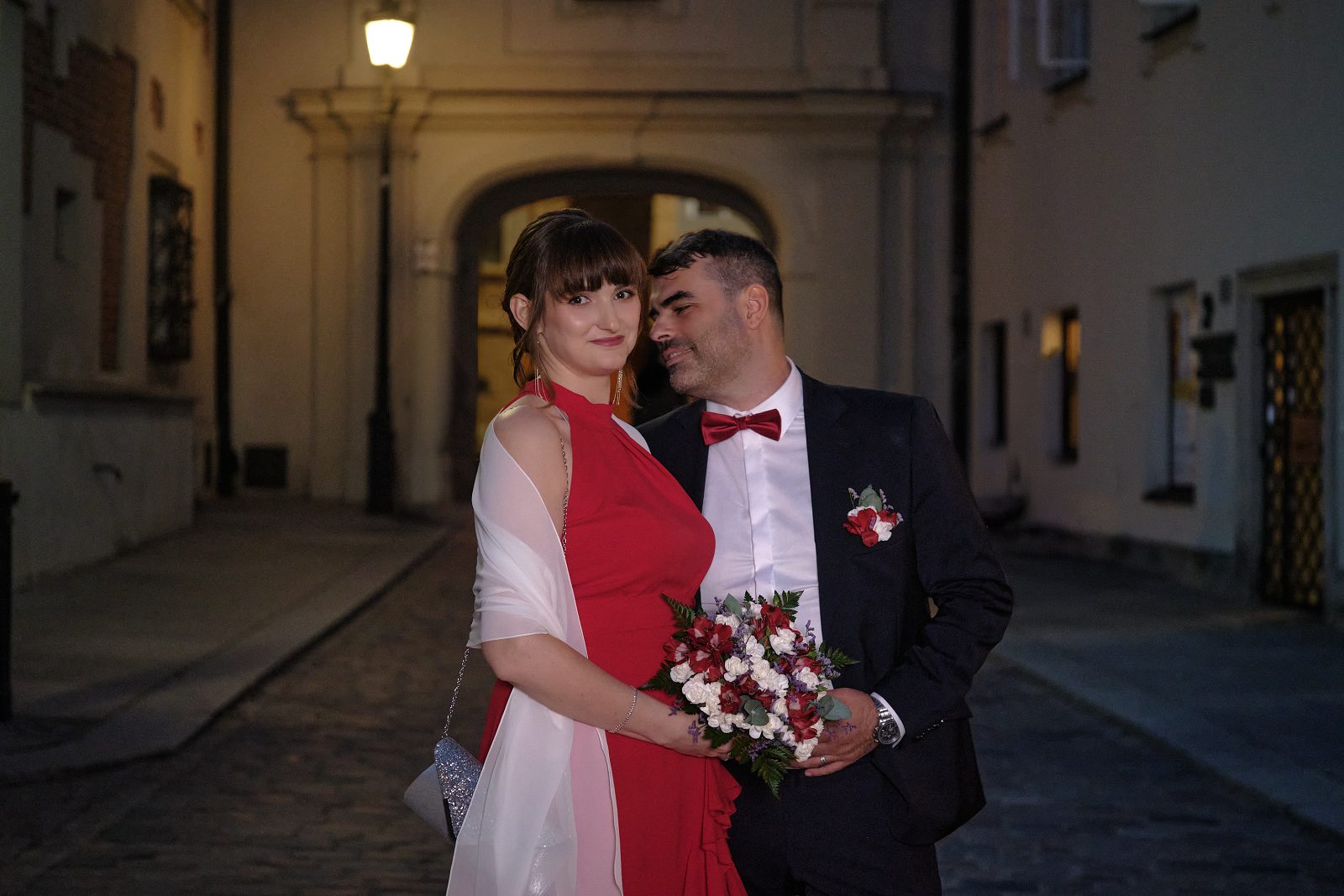Pan młody z bukietem pochyla się ku pannie młodej na nocnej ulicy.