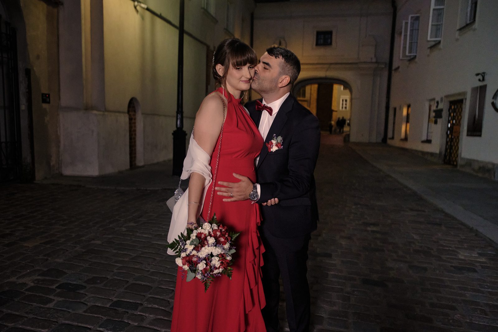 Pan młody całuje pannę młodą w policzek; ona trzyma bukiet.