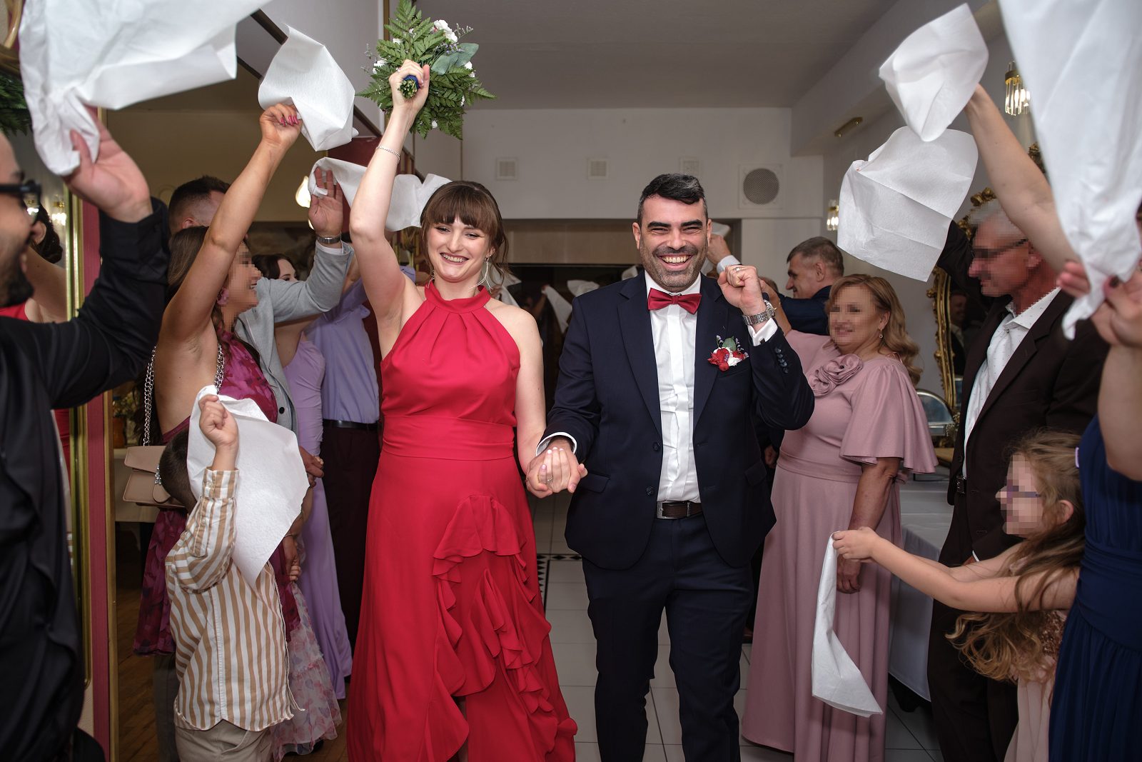 Nowożeńcy przechodzą przez restaurację wśród gości unoszących białe serwetki.